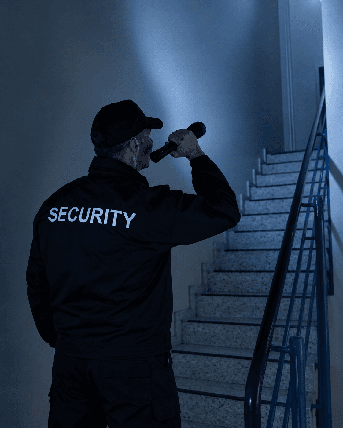 Security guard in black uniform shining a flashlight up a dark stairwell during a night patrol.