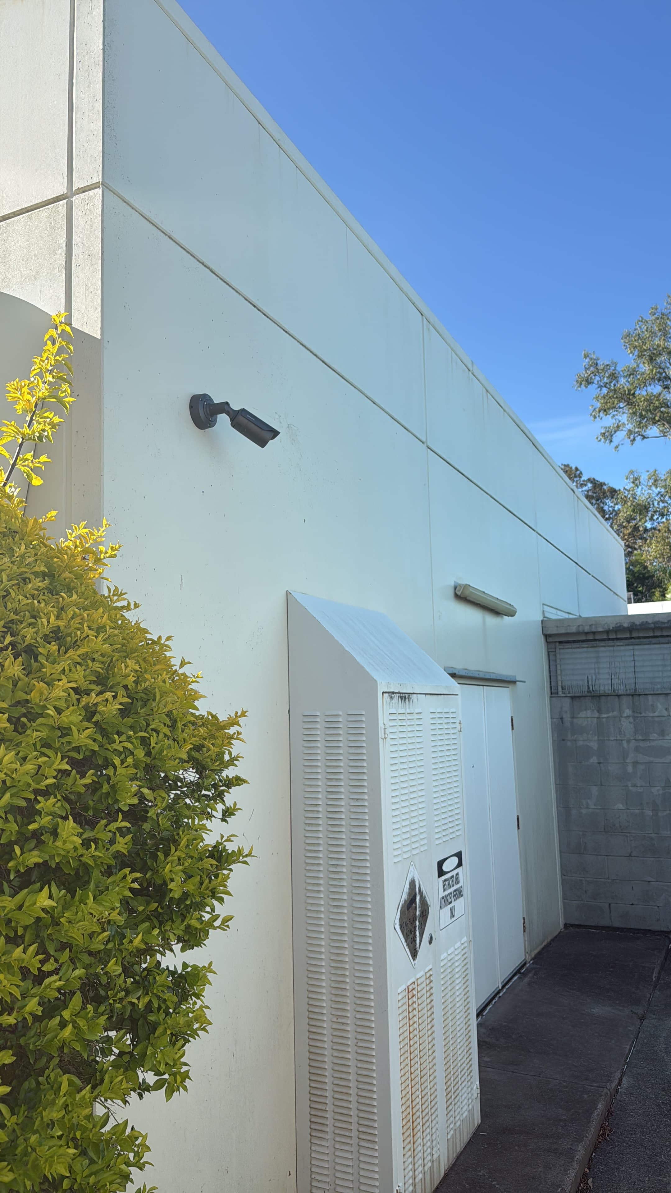 Security camera mounted on a white wall above a large utility box and green shrubbery.