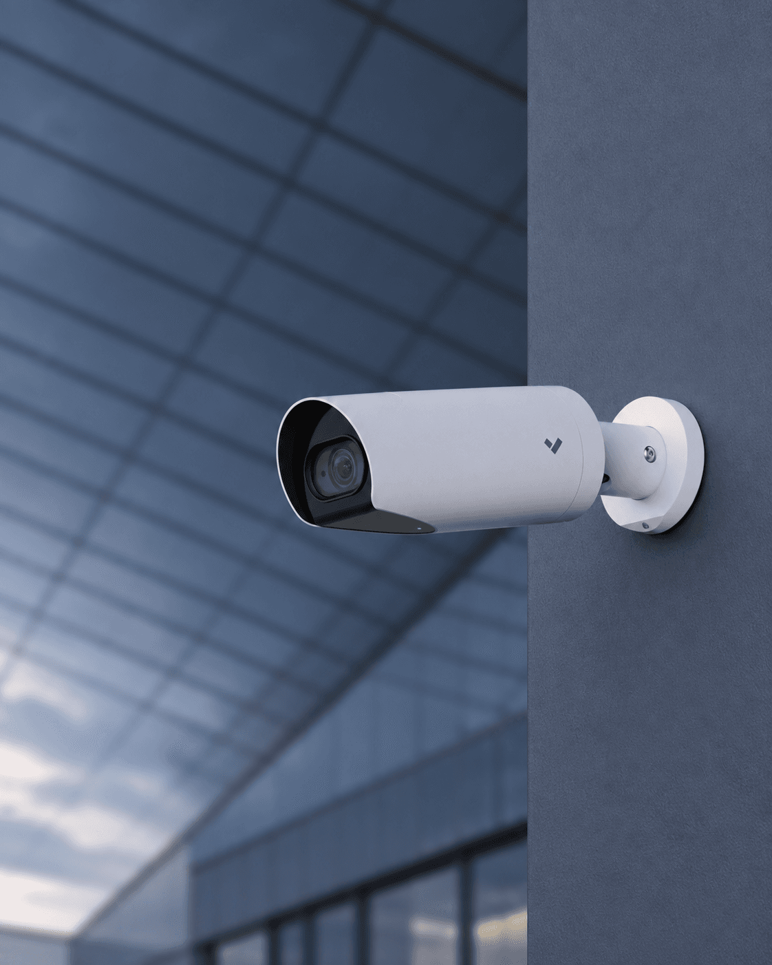 White bullet security camera mounted on a dark wall outside a modern glass building.