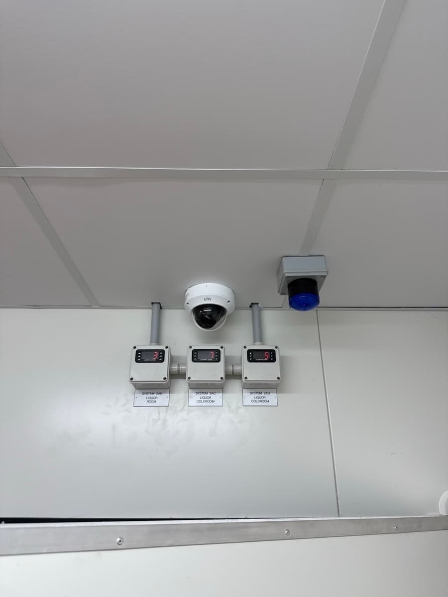 Three digital temperature displays for liquor coldrooms below a security camera and blue alarm light.