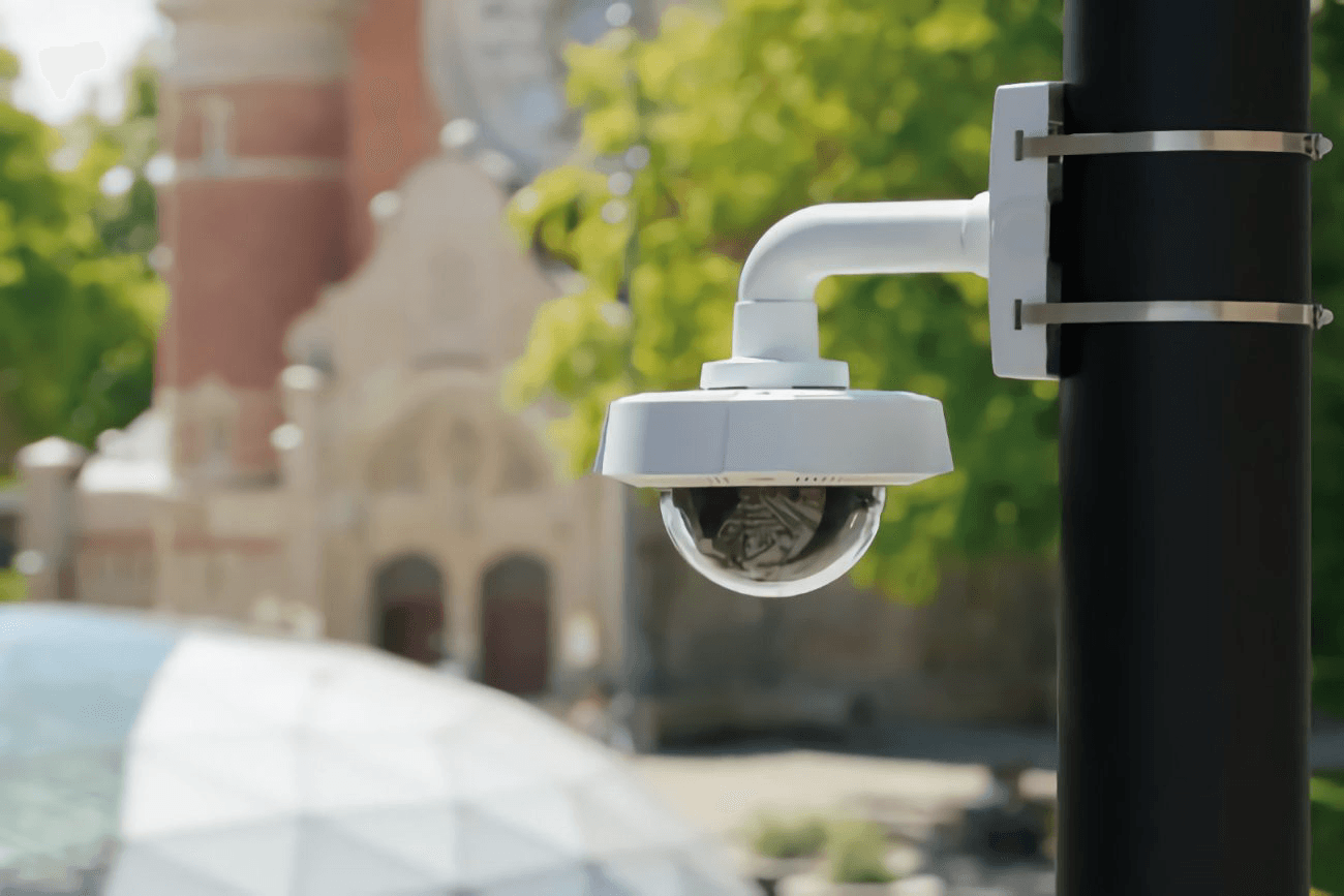 White dome security camera mounted on a black pole with a blurred campus background.