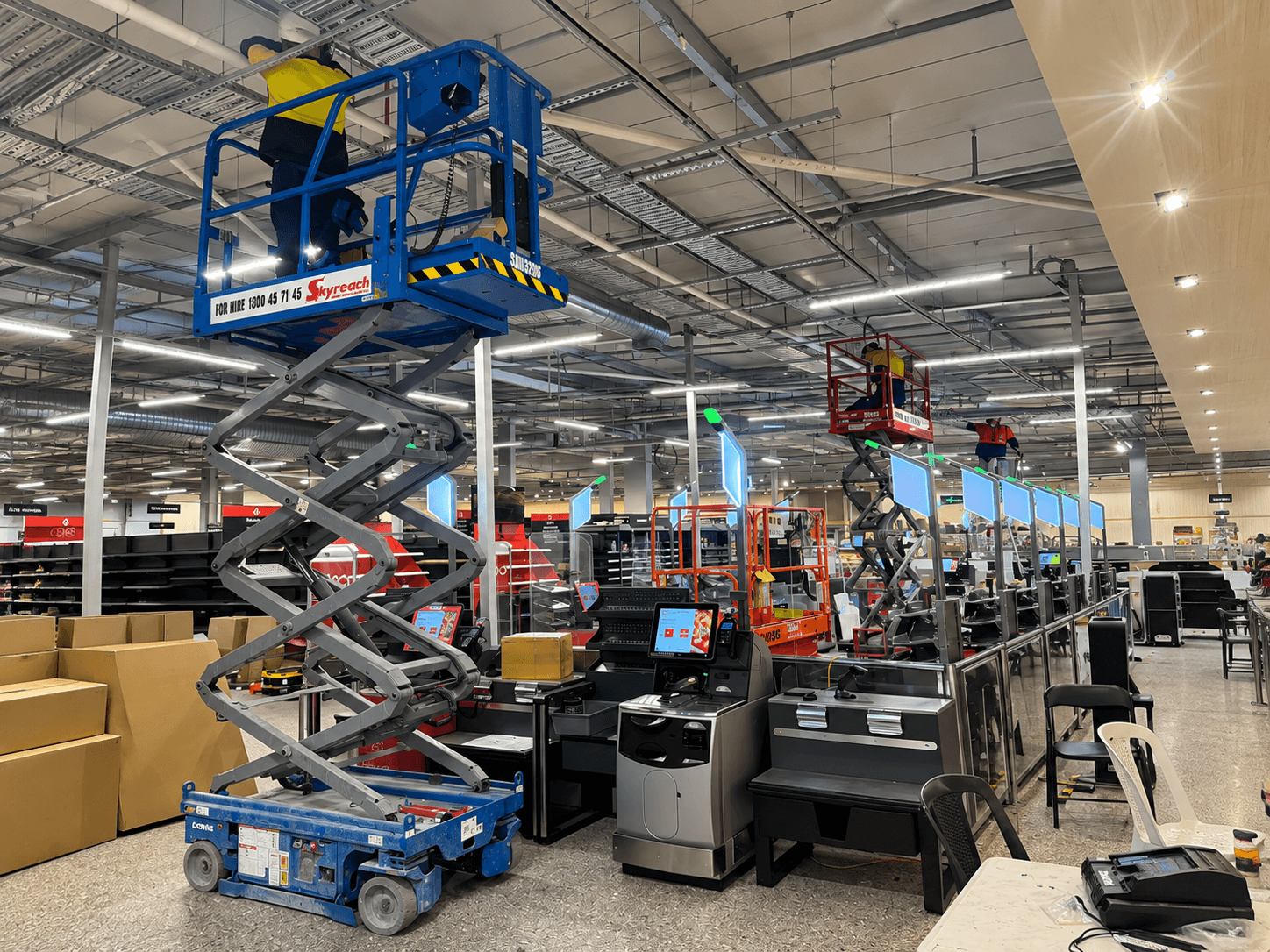 Workers on scissor lifts perform ceiling maintenance inside a large retail store under construction.