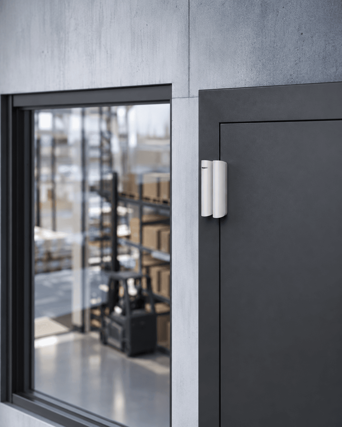 White magnetic door sensor on a dark door beside a window overlooking a warehouse interior.