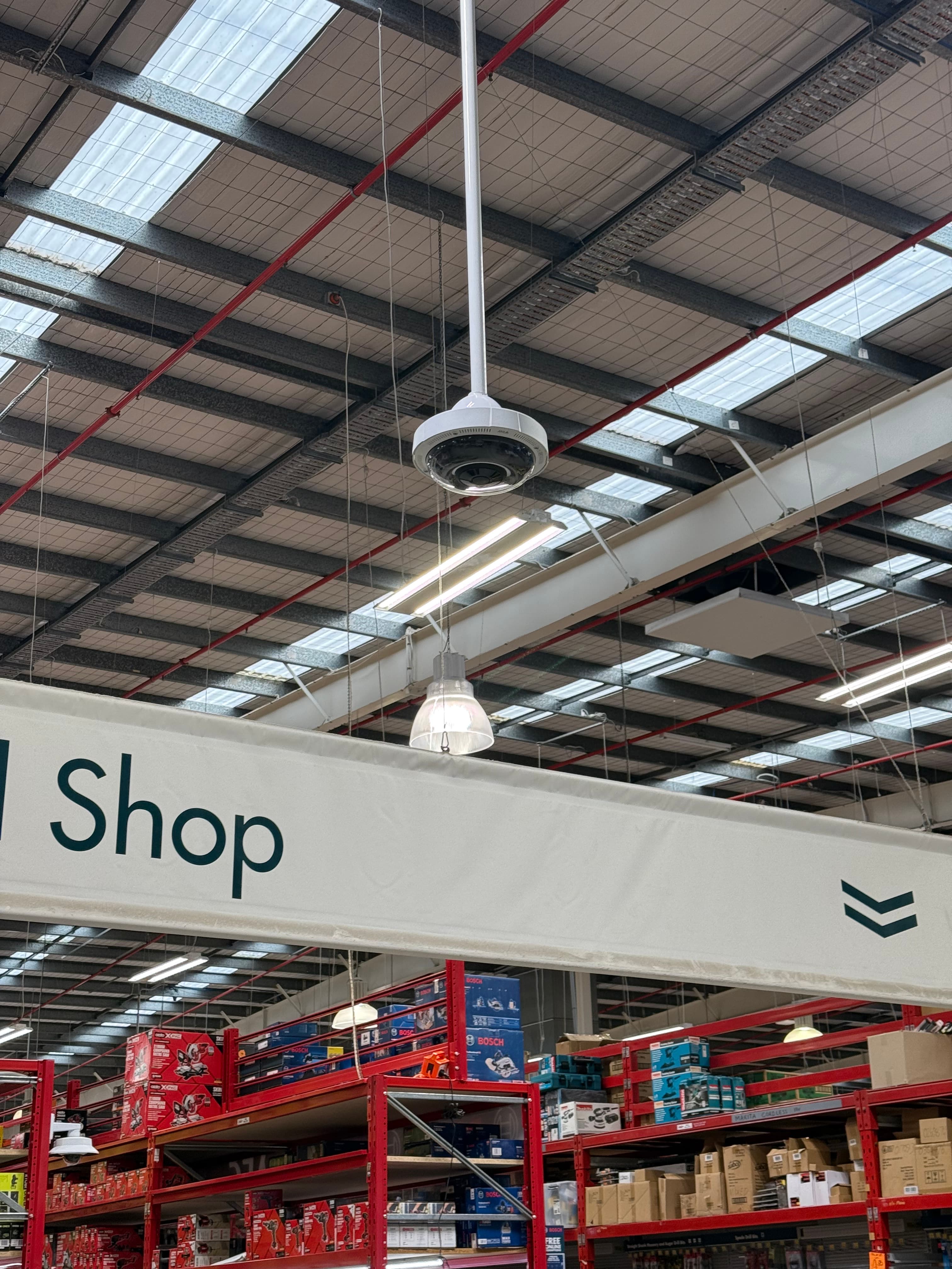 Security camera and shop banner suspended from a high warehouse ceiling above stocked tool shelves.