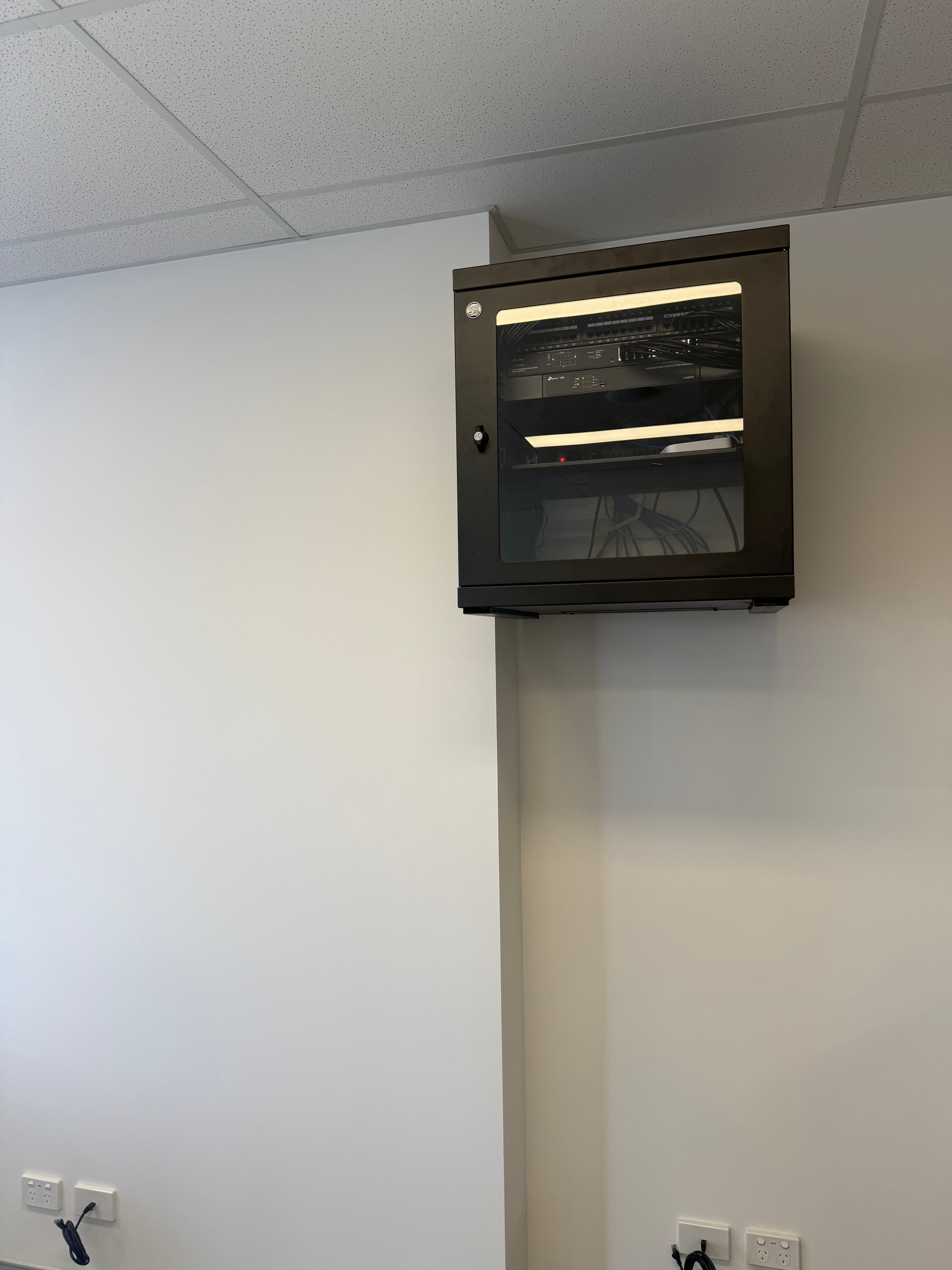 Black wall-mounted network cabinet with glass door housing networking equipment on a white wall.