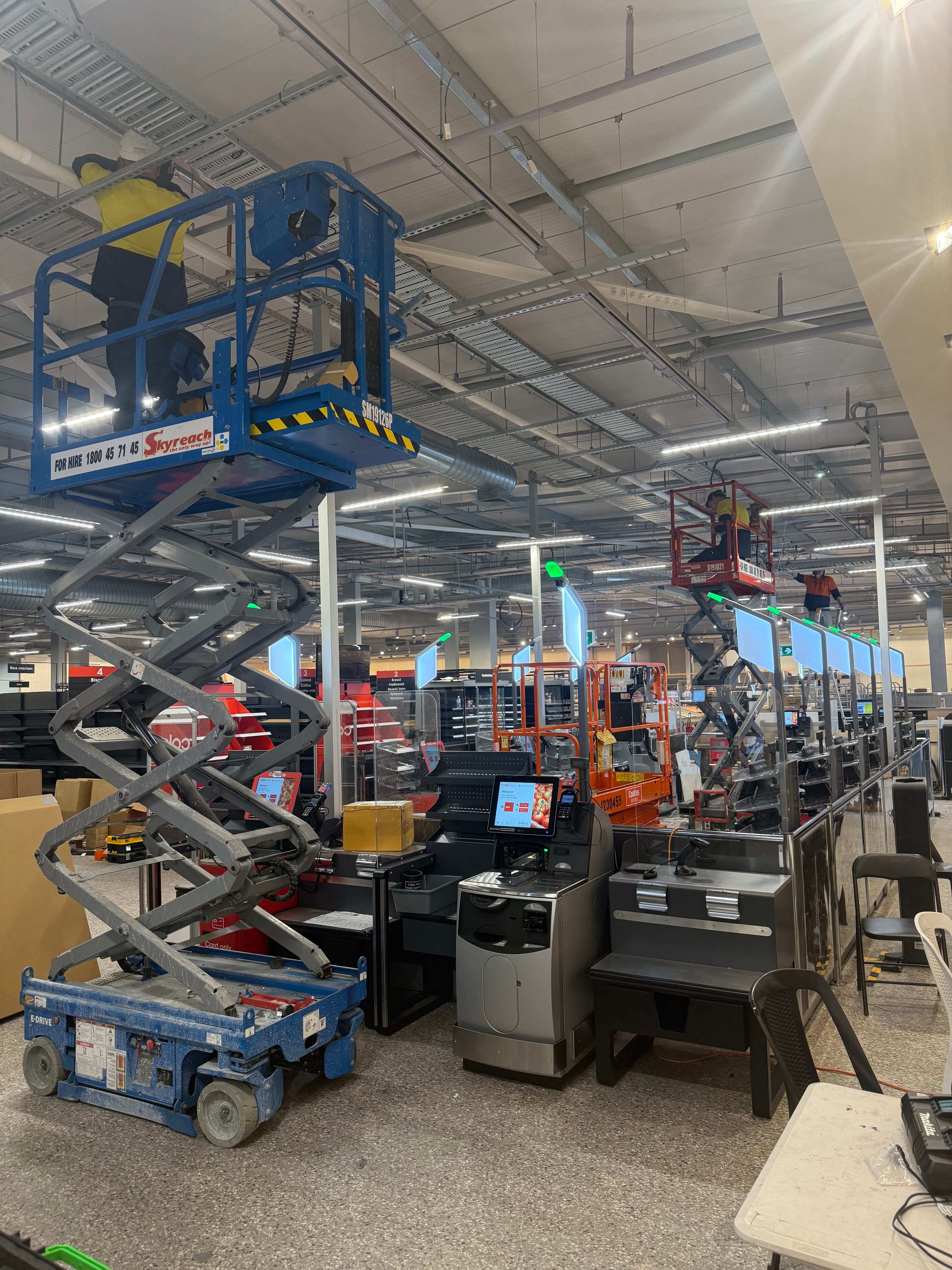 Workers on blue and orange scissor lifts perform maintenance in a large retail store.