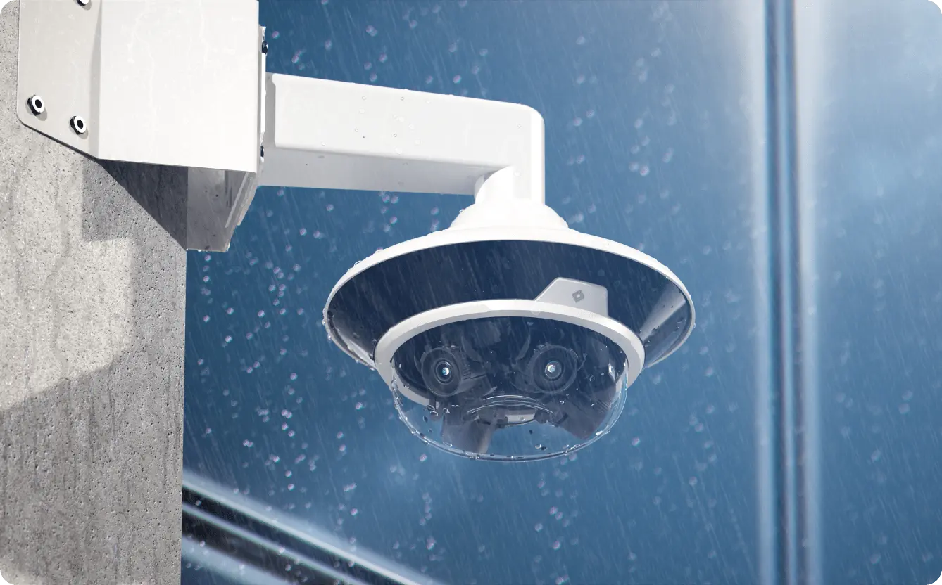 White multi-sensor security camera mounted on a concrete wall during a heavy rainstorm.