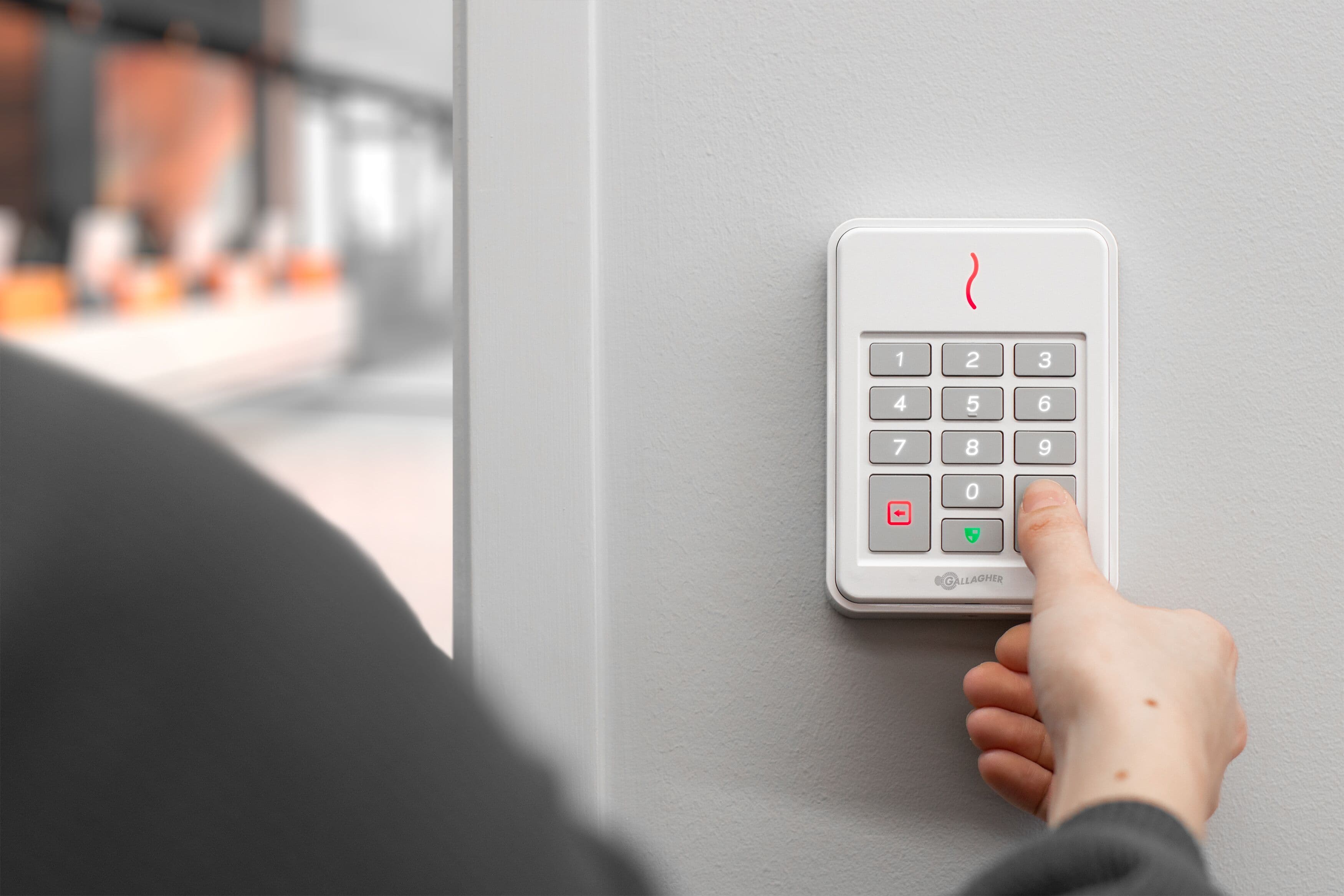 Hand pressing a button on a white Gallagher security keypad mounted on a grey wall.