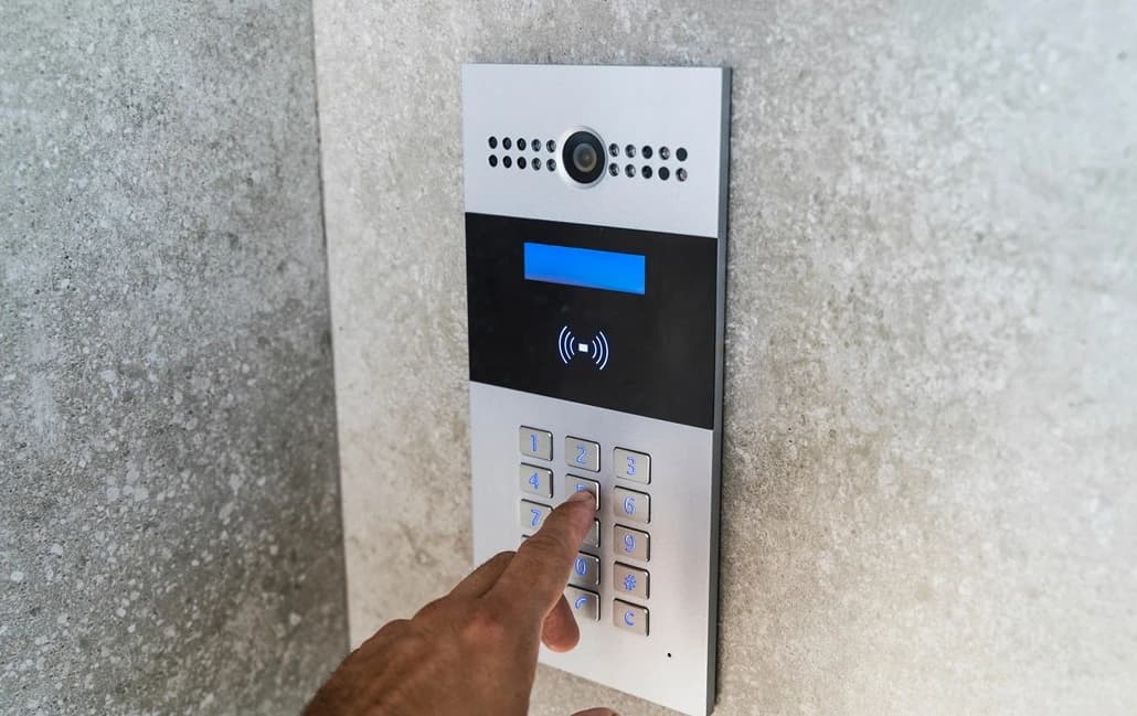 Hand pressing a button on a silver intercom with a camera and numeric keypad.