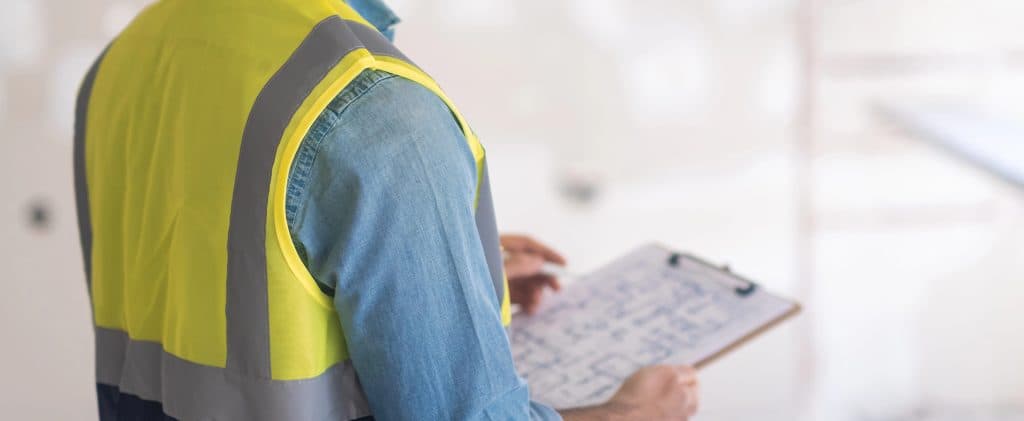 Construction worker in a yellow safety vest holding a clipboard with blueprints at a site.