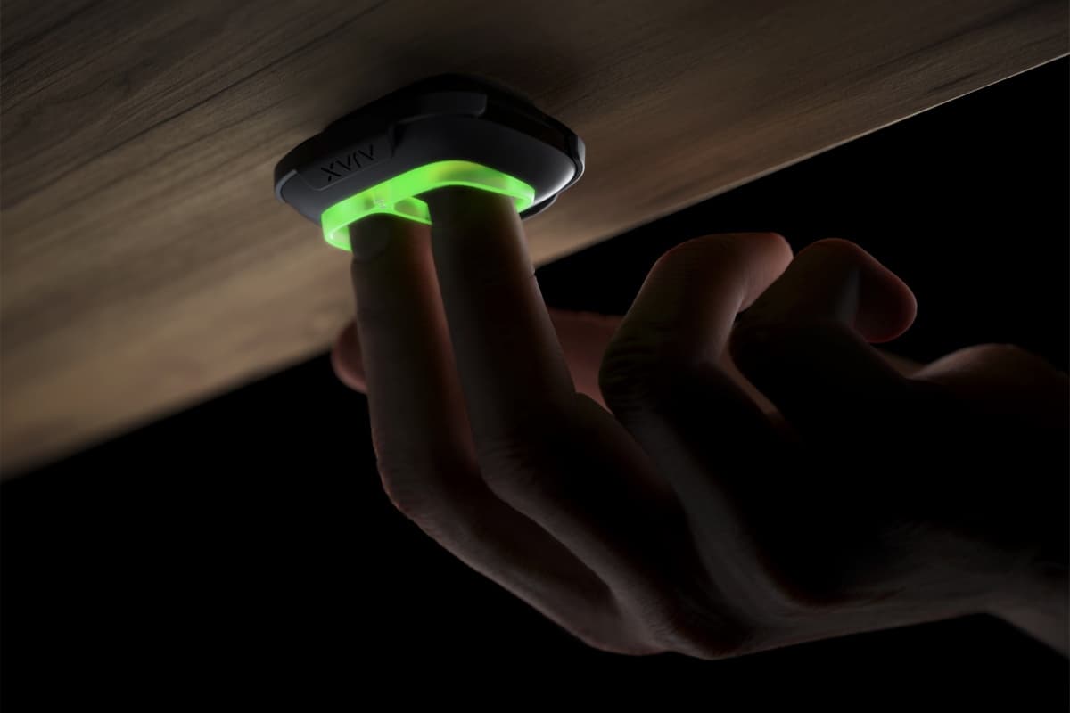 Hand pressing a glowing green Ajax panic button mounted under a wooden desk surface.