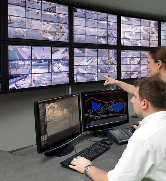 Security personnel monitor a wall of surveillance feeds and computer screens in a control room.