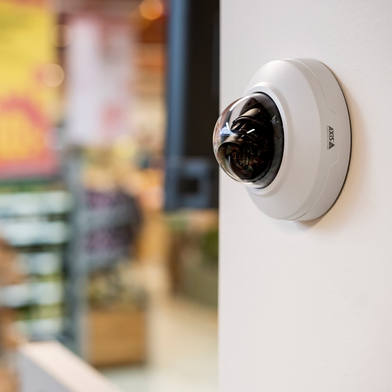 White dome security camera mounted on a wall overlooking a blurred retail store interior.